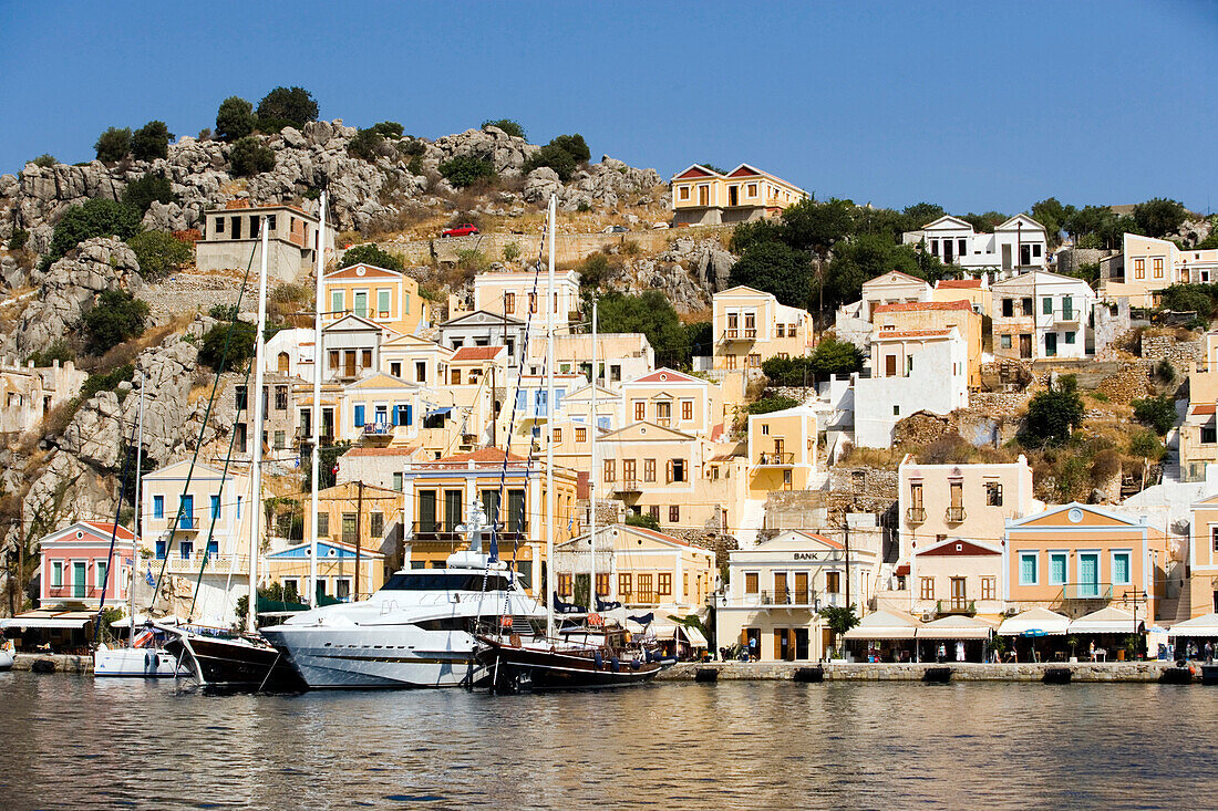 Yacht and sailing boats anchoring at quay of harbour Gialos, picturesque mansions at mountainside in background, Simi, Symi Island, Greece