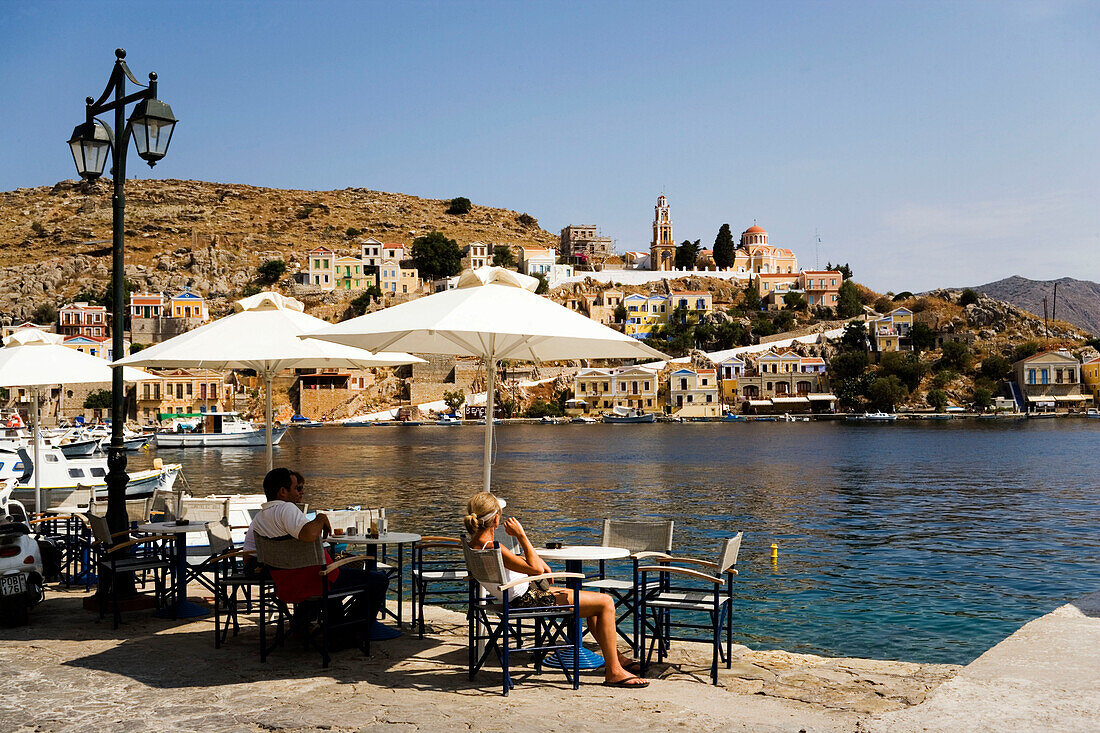 People sitting in pavement cafe and looking over the harbour Gialos to monastery Moni Evangelismos, Simi, Symi Island, Greece