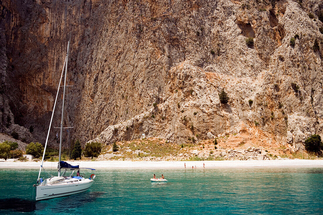 Sailing boat anchoring near beach, people at beach of Sisalona Bay, rock face in background, Symi Island, Pedi, Greece