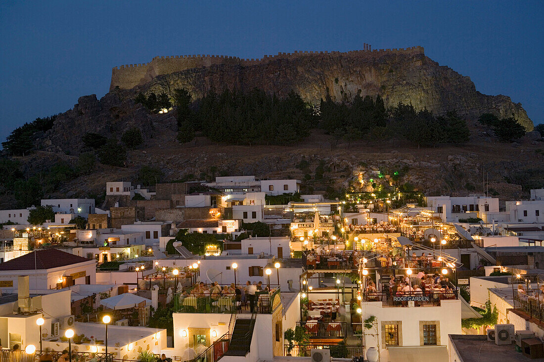 View over illuminated town in the evening to Acropolis, people sitting on terraces of restaurants, Lindos, Rhodes, Greece