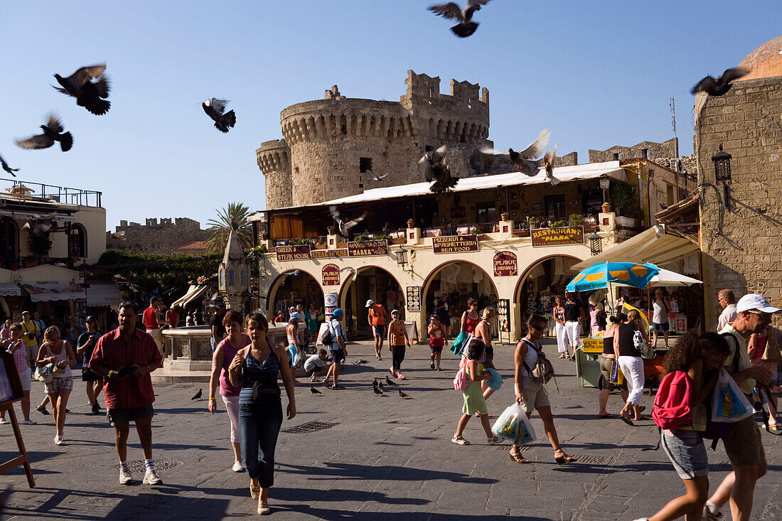 People strolling over busy Platia Ippokratou with shops and restaurants, flying doves, Thalassini Gate in background, Rhodes Town, Rhodes, Greece, (Since 1988 part of the UNESCO World Heritage Site)