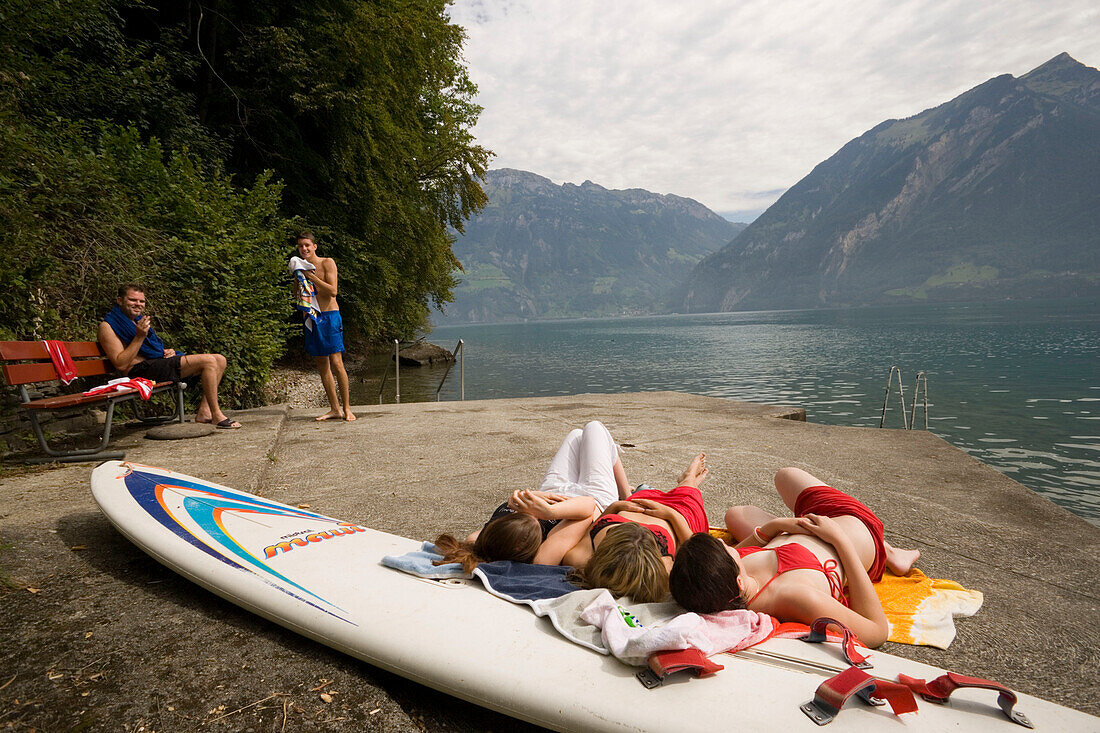Three girls sunbathing on rock at lake Urnersee, boys watching them, Bauen, Canton of Uri, Switzerland
