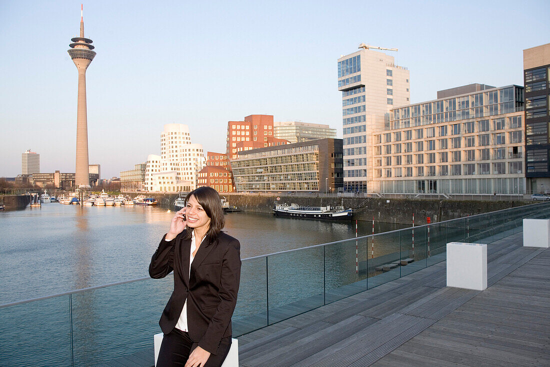 Young business woman talking on a mobile phone, in front of the city skyline, television tower, Zollhof, Media Harbour, architecture of Frank O.Gehry, Düsseldorf, state capital of NRW, North-Rhine-Westphalia, Germany