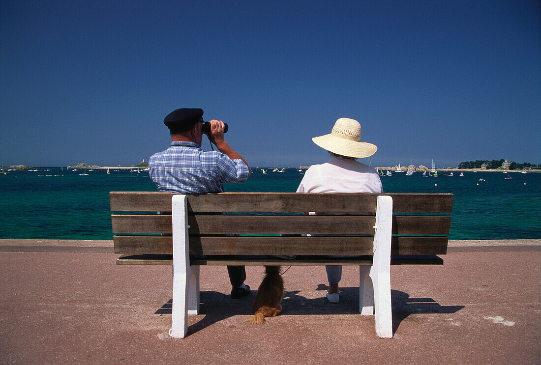 Two people sitting on a bench, man using binoculars, Port Blanc, Brittany, France