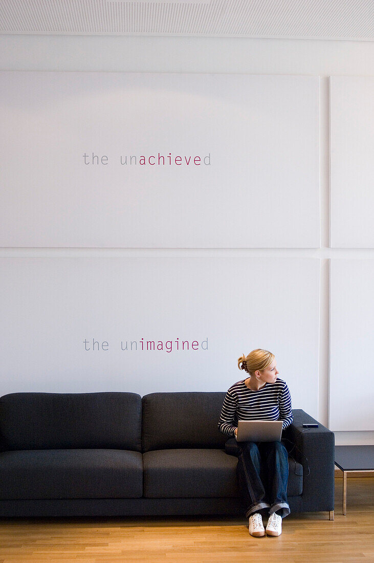 Junge Frau mit Laptop sitzt auf einem Sofa und blickt zur Seite, Luxemburg