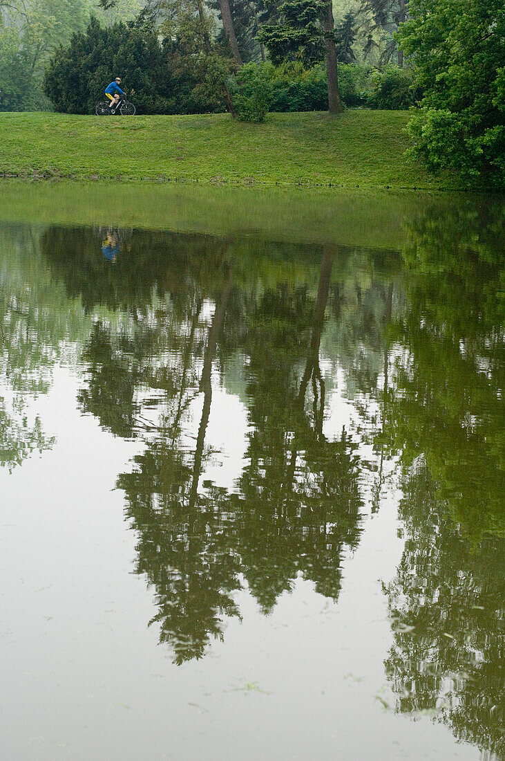Bäume und ein Radfahrer spiegeln sich im ruhigen Wasser eines Sees, Lazieni Park, Warschau, Polen