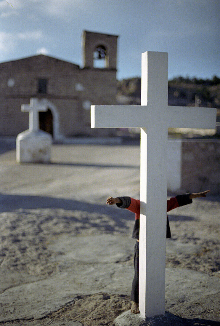 White cross in front of church, a child is hiding behindthe cross, Creel, Mexico