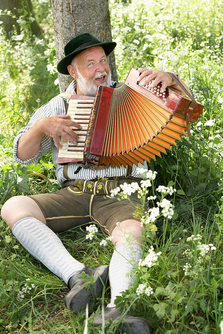 Man wearing bavarian costume leaning against a tree while playing melodeon