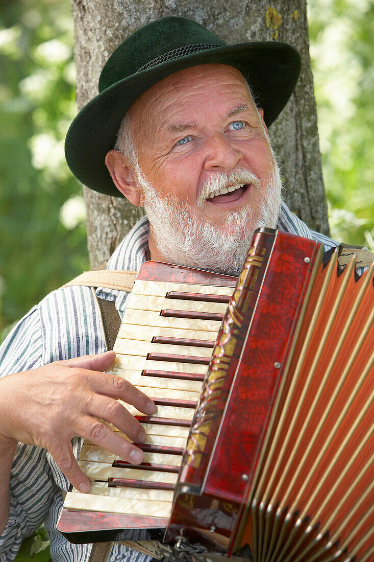Mann in bayrischer Tracht lehnt gegen einem Baum und spielt Akkordeon