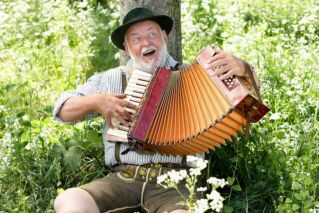 Man wearing bavarian costume leaning against a tree while playing melodeon