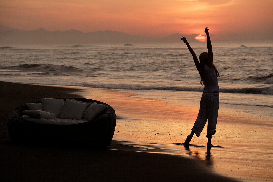 Eine junge Frau dehnt sich am Strand bei Sonnenuntergang, nahe Uluwatu, Bali, Indonesien