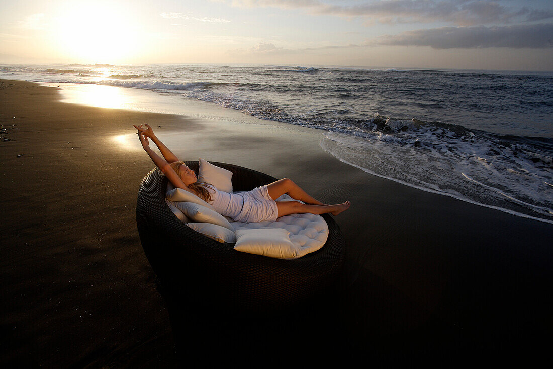 Eine junge Frau entspannt sich am Strand bei Sonnenuntergang, nahe Uluwatu, Bali, Indonesien