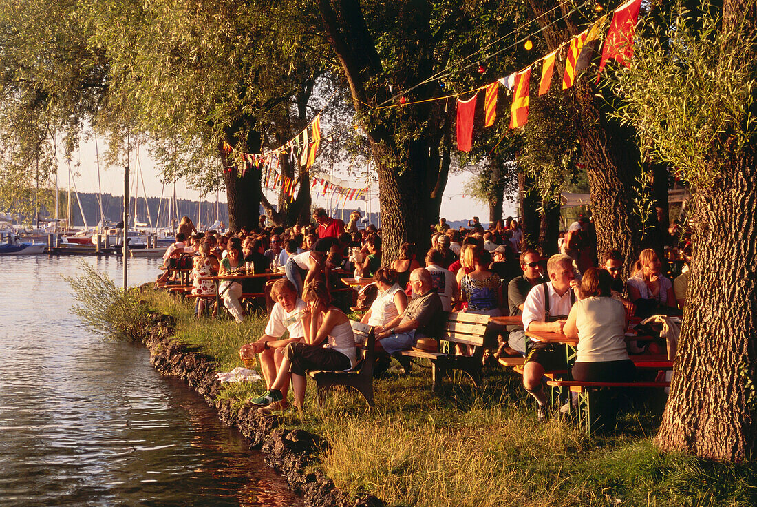 Frauenchiemsee, island feast in July, people sitting on benches under trees by the lake Chiemsee, Chiemgau, Upper Bavaria, Germany