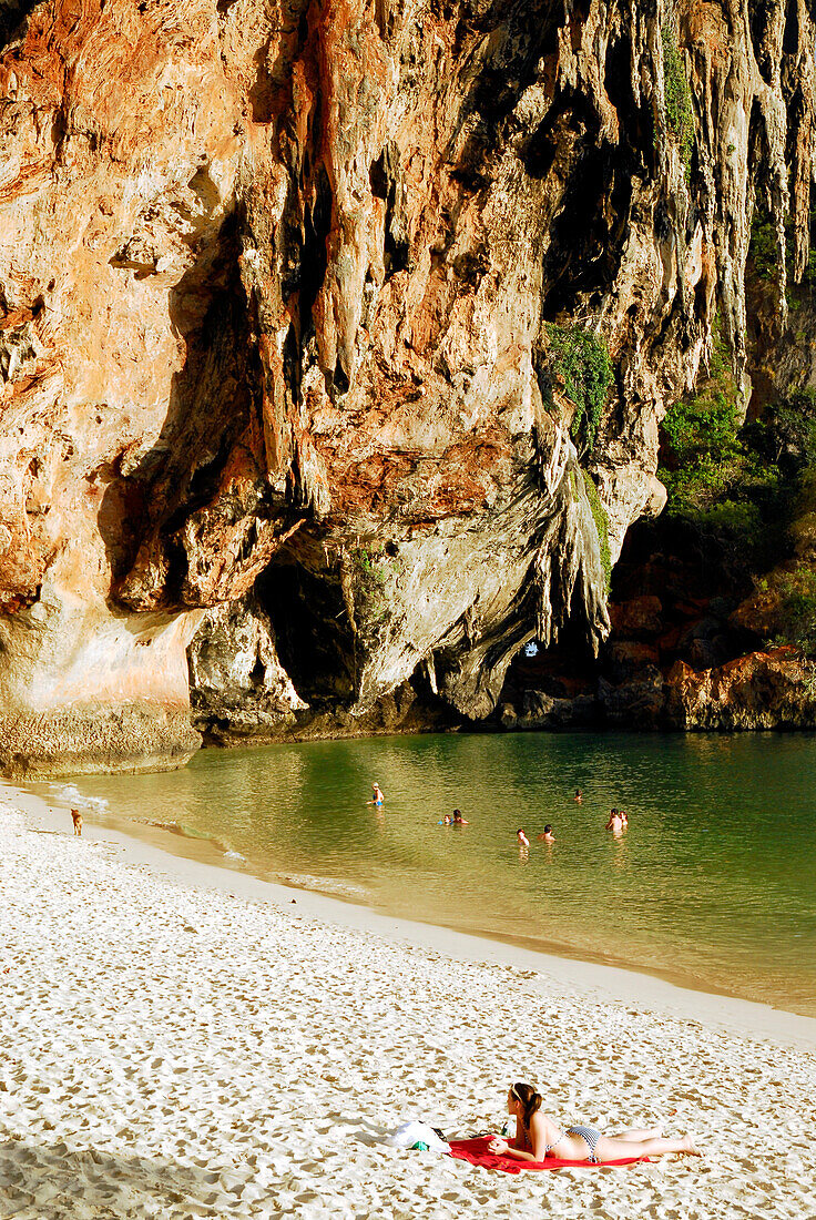 People sunbathing and swimming in the water near limestone cliffs at Hat Phra Nang beach, Krabi, Thailand