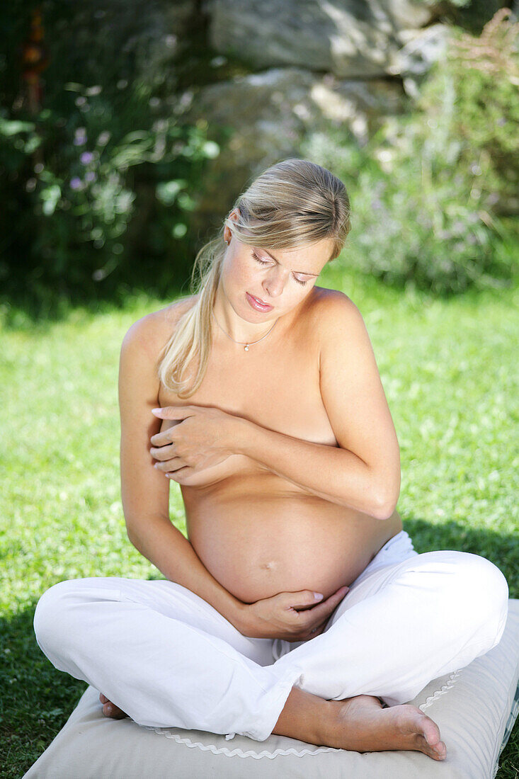 Schwangere Frau sitzt mit freiem Oberkörper im Garten, Steiermark, Österreich