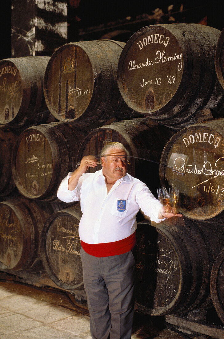 A wine taster pouring samples of Sherry into tasting glasses in front of a heap of Sherry casks, in the Bodegas Pedro Domeq, Jerez de la Frontera, Cadiz province, Andalusia, Spain