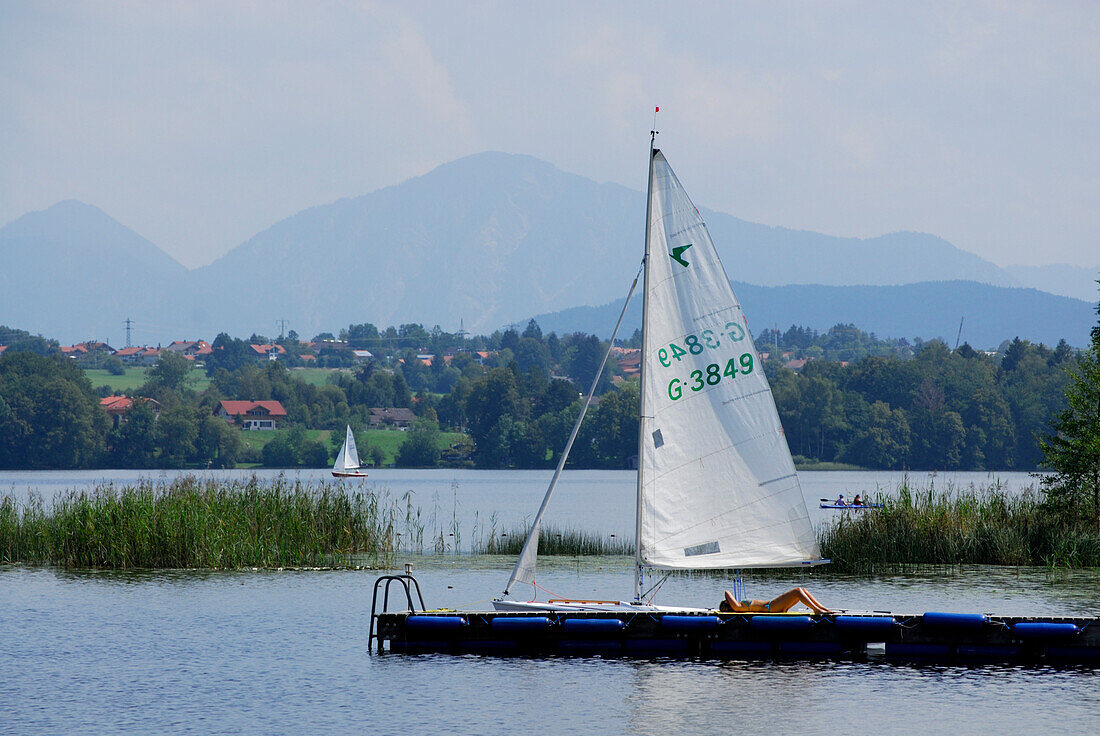 young woman sunbathing on landing stage with sailing boat, Bavarian Alps in background, lake Staffelsee, Osterseen, Upper Bavaria, Bavaria, Germany
