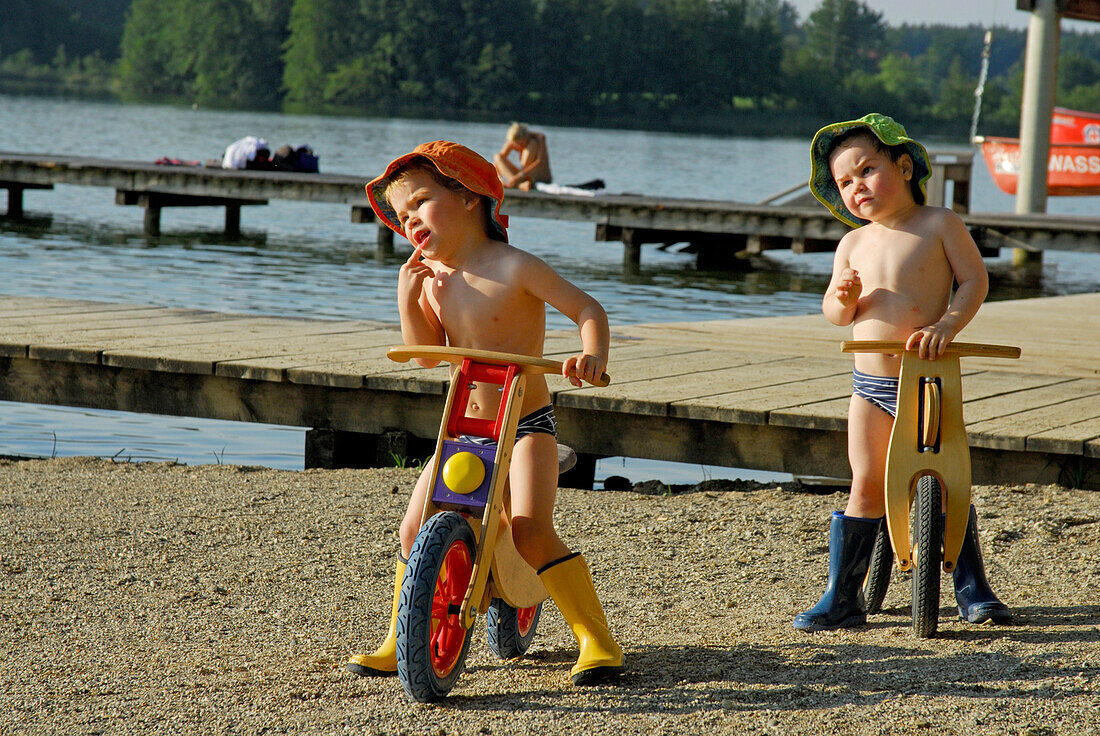 two boys with sunhat on juvenile bicycle, beach of lake Hartsee, Chiemgau, Upper Bavaria, Bavaria, Germany