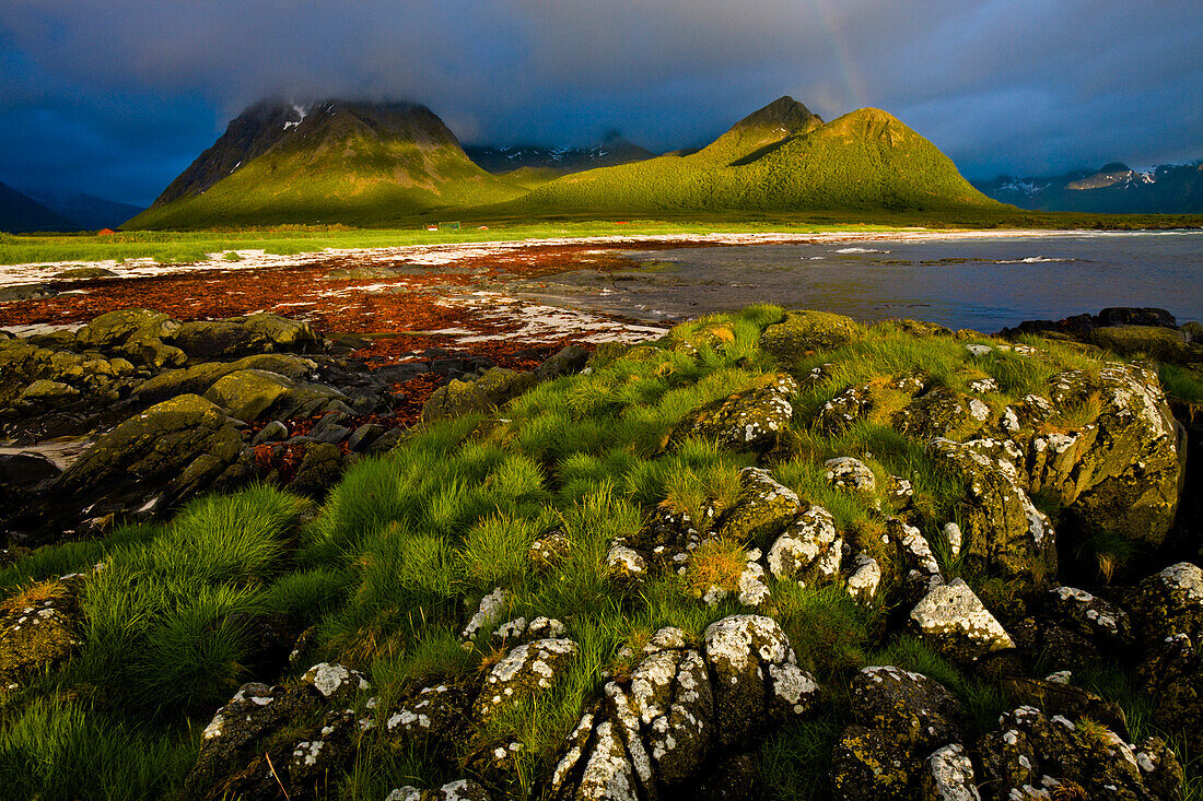 Mitternachtsonne und Wolken verhangene Berge am Strand von Hadselsand, Ein Regenbogen bildet sich in den dunklen Wolken, Insel Austvagoya, Lofoten, Norwegen.
