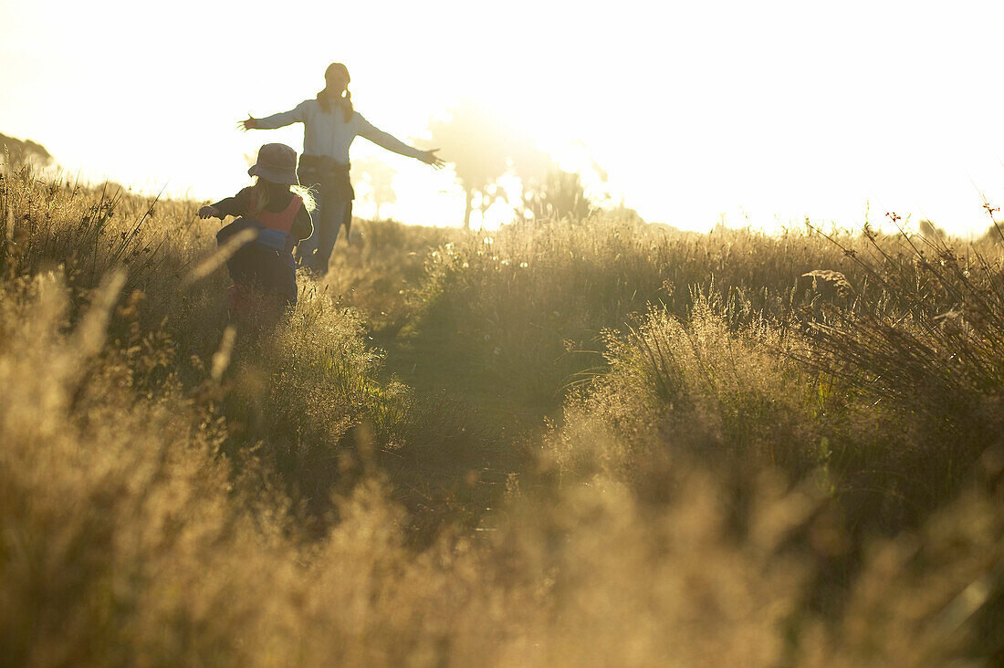 Mutter und Kind laufen durch hohes Gras, Sonnenuntergang an der Westküste, bei Haast, Südinsel, Neuseeland