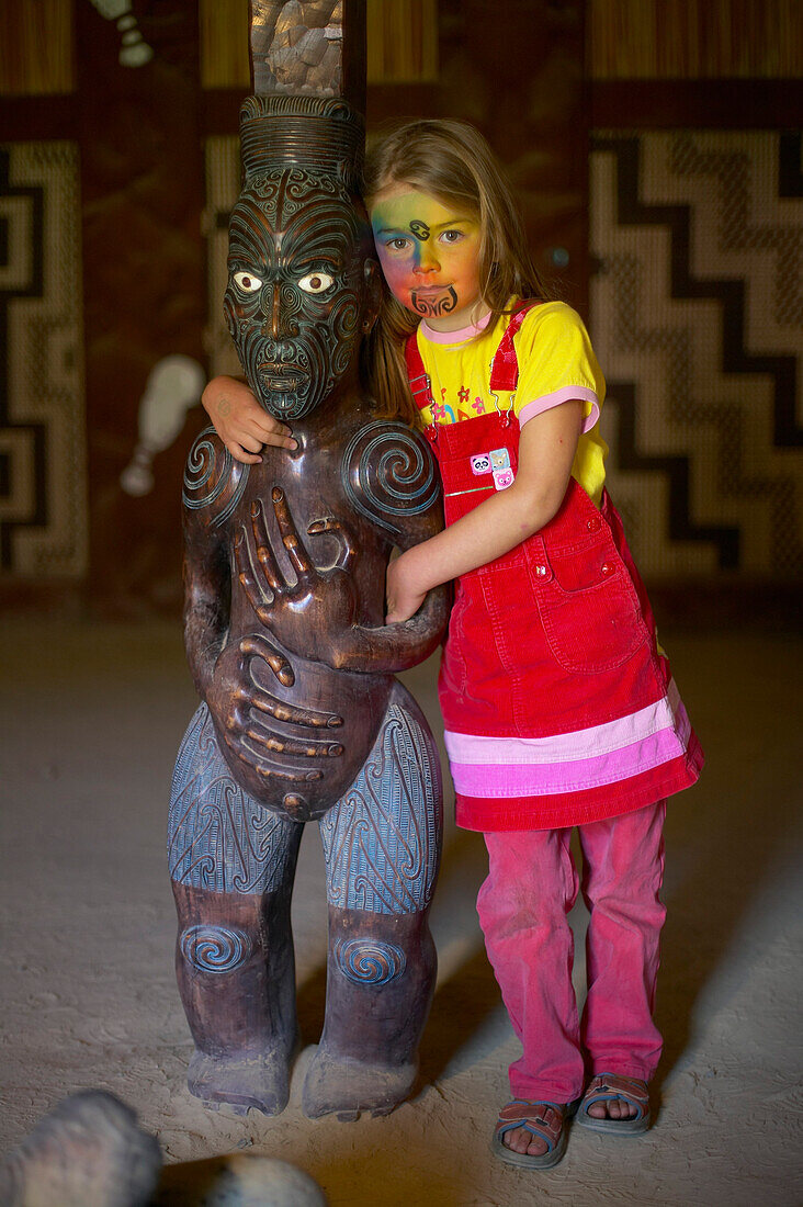 Mädchen mit Facepainting, nachempfundenes Moko (Maori Gesichtstatoo), geschnitzte Maori Figur in Versammlungshaus (Marae),  Okains Bay, Bank's Peninsula, Ostküste, Südinsel, Neuseeland
