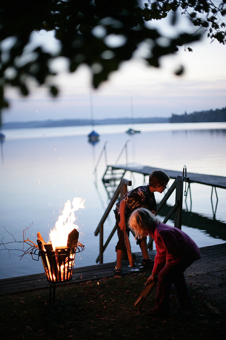 Two children standing around a fire near the jetty, evening, Lake Woerthsee, Upper Bavaria, Bavaria, Germany