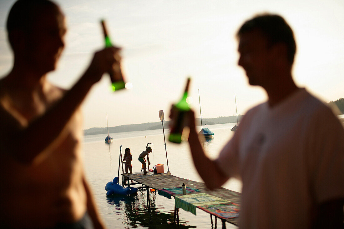 Two men drinking bottles of beer, children playing on jetty in background, Lake Woerthsee, Bavaria, Germany, MR