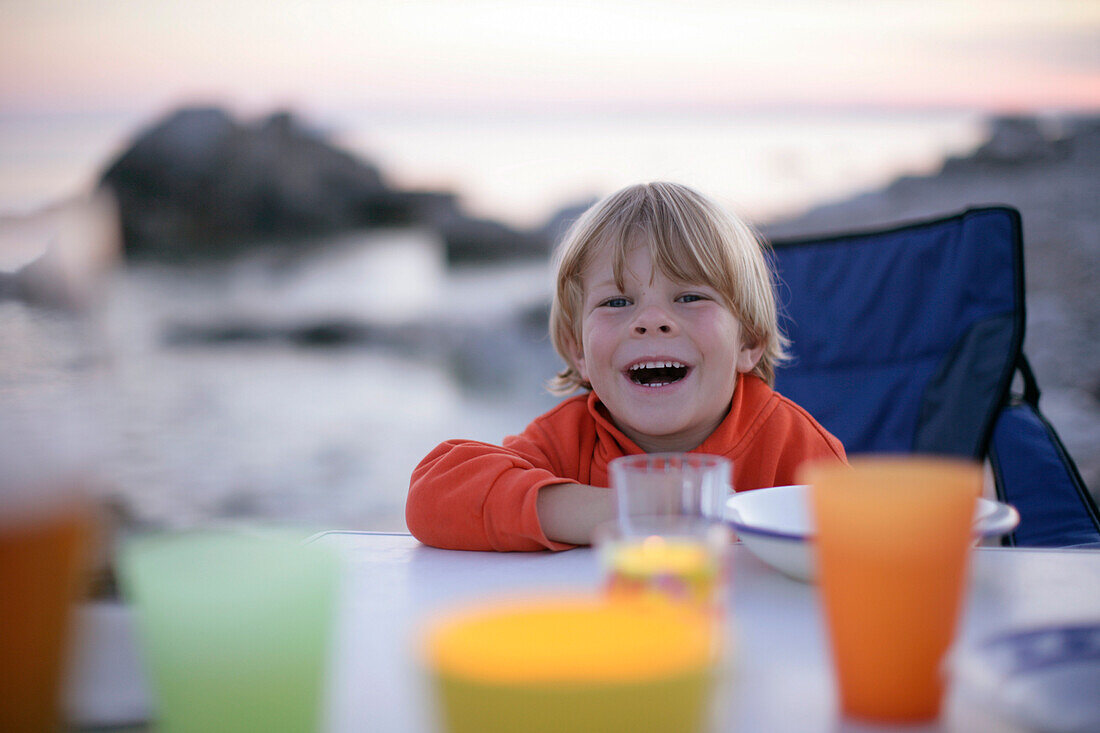 Junge sitzt am Abendtisch und lächelt, Meer im Hintergrund, Sysne, Gotland, Schweden