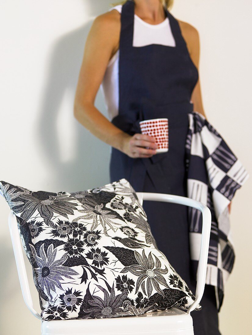 Pillow with black and white design on a chair and a woman with a black apron and cup in her hand