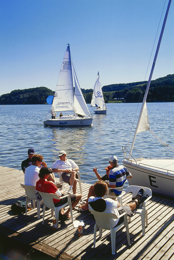 A group of people sitting on a jetty at a lake, sailing boats in the background, Baldeney lake, Essen, Ruhr Valley, Ruhr, Northrhine, Westphalia, Germany