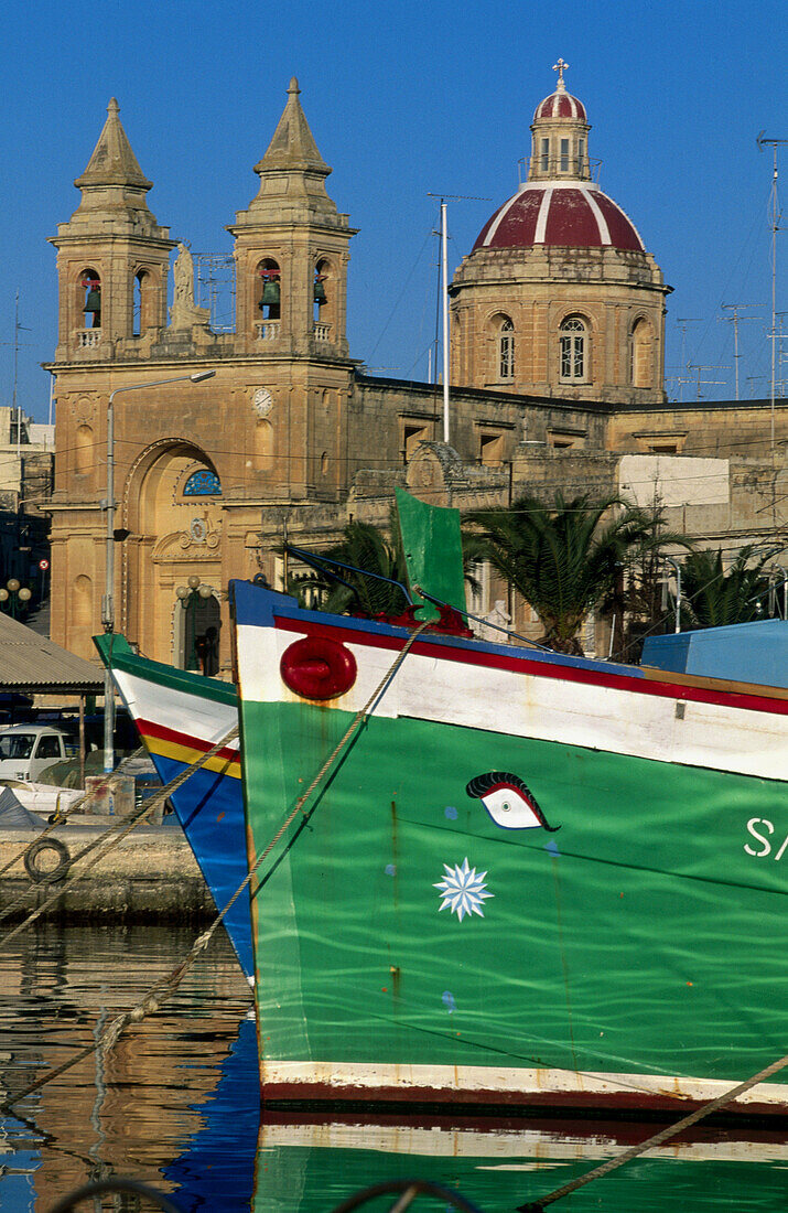The church and fishing boats Luzzu . Marsaxlokk fishermen harbour. Republic of Malta.