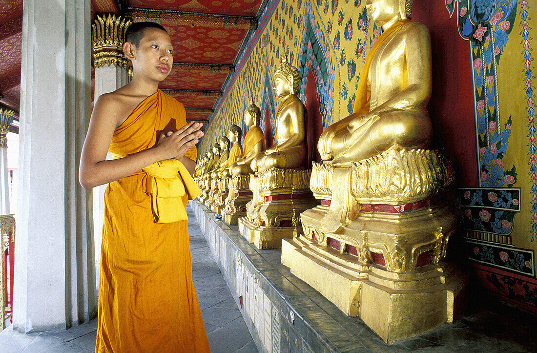 Young buddhist novice monk at prayes in the Buddhas statues lining the cloister. Wat Arun Temple. Bangkok. Thailand