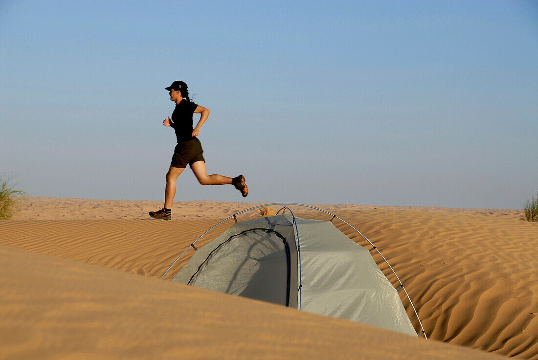 Woman jogging over dune, tent in foreground, Djebel Tembaine, Tunisia