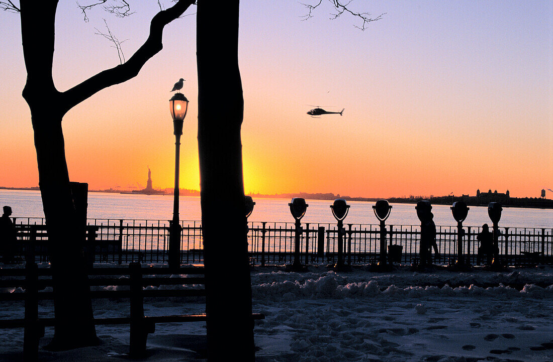 Sunset view from Battery Park, New York City, showing water and a helicopter silhouetted in the sky and trees, street light and coin operated binoculars silhouetted in the foreground