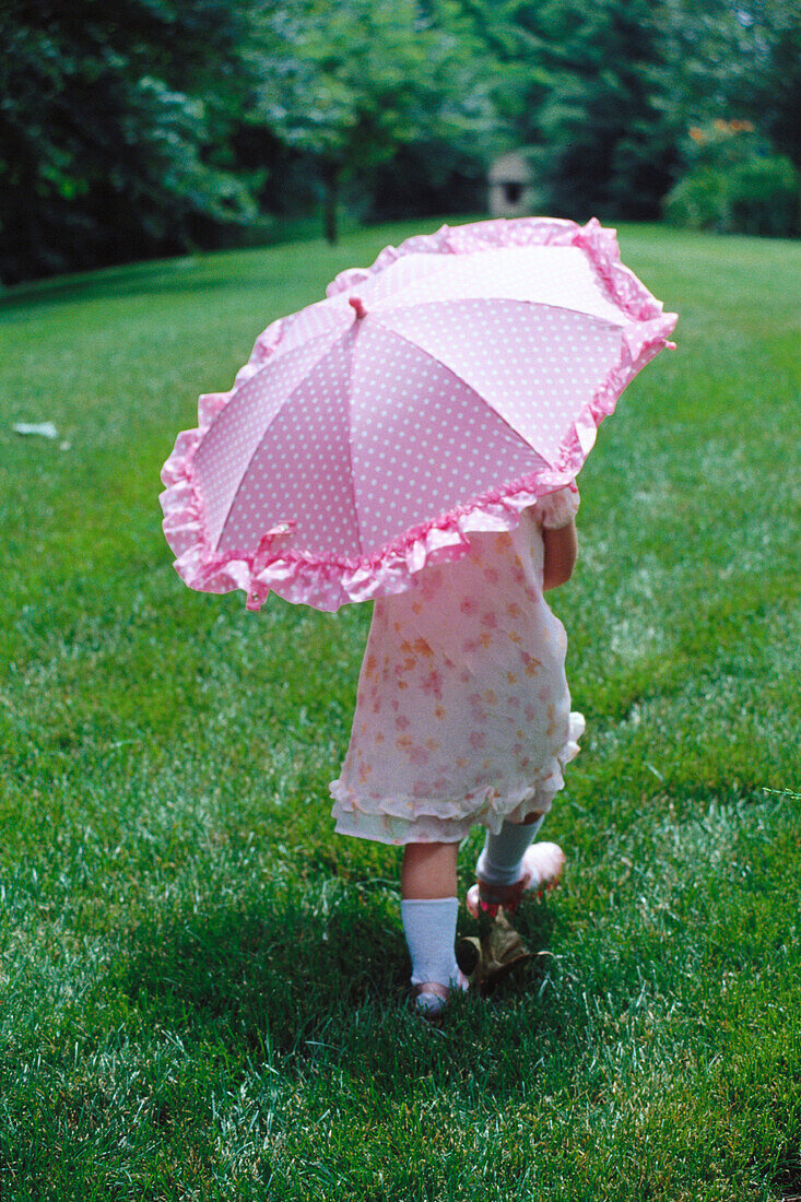 Small girl walking, holding a pink umbrella, viewed from behind