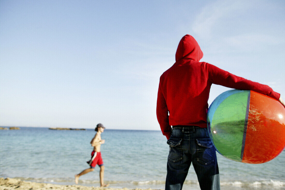  Adult, Adults, Back view, Ball, Balls, Beach, Beach ball, Beach balls, Beaches, Coast, Coastal, Color, Colour, Contemporary, Contrast, Contrasts, Daytime, Exterior, Fit, Hold, Holding, Hood, Hoods, Human, Knees-up, Leisure, Outdoor, Outdoors, Outside, Pa