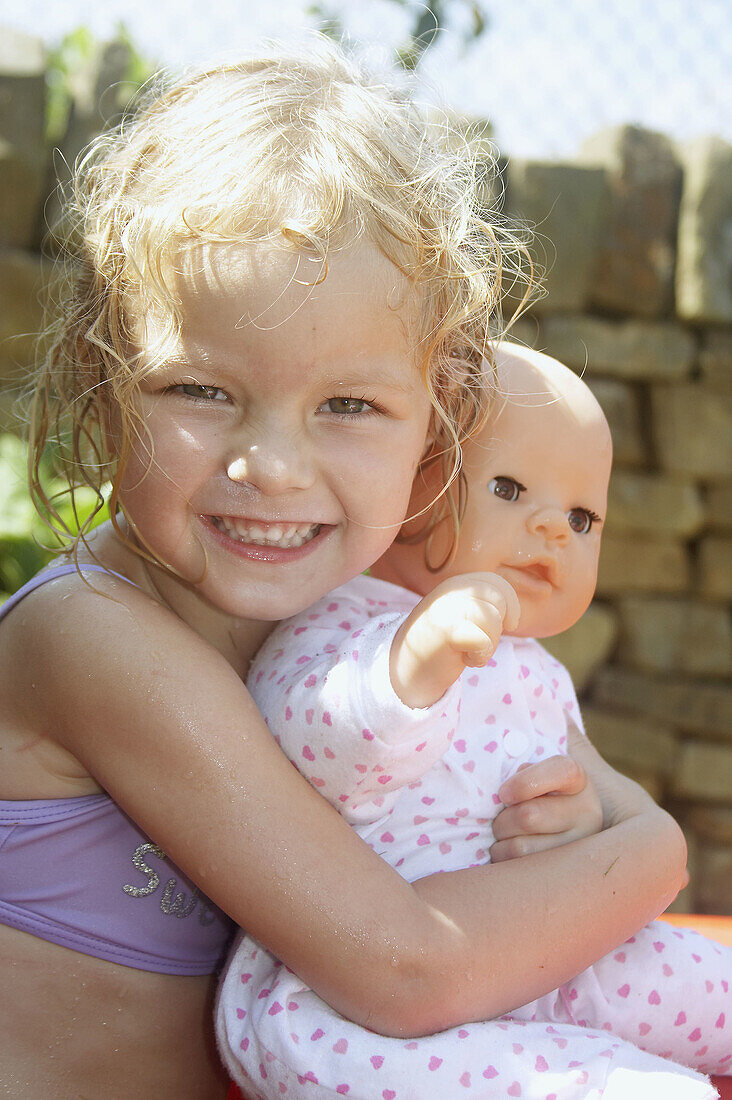 3 year old girl outside in swimming costume holding a doll smiling into camera