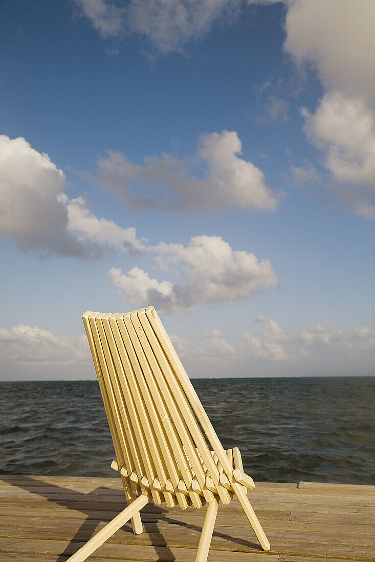 BELIZE Ambergris Caye Slatted yellow wooden chair sit at end of wooden dock, partly cloudy skies, Caribbean waters to horizon