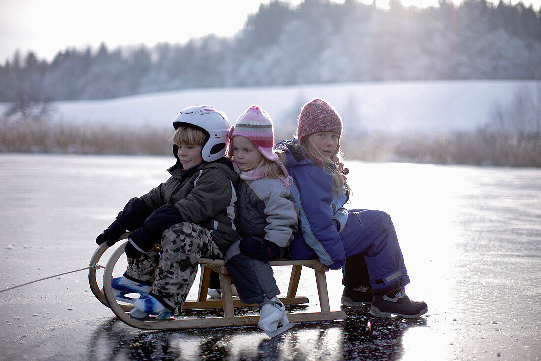 Children sitting on sledge on frozen lake Buchsee, Munsing, Bavaria, Germany
