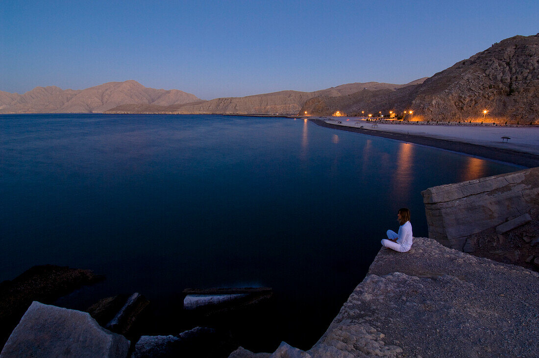 Frau sitzt auf Felsen und betrachtet den Aussicht, Küstenlandschaft und Strand im Abendlicht, Kashab, Khasab, Musandam, Oman