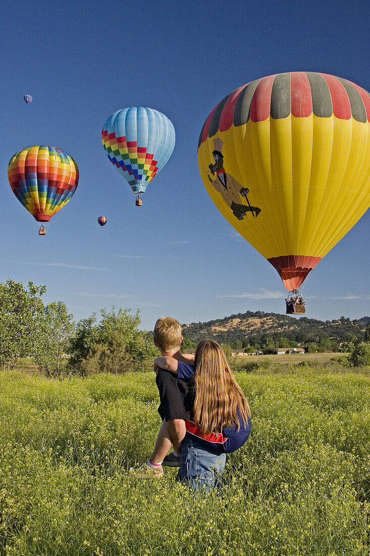A girl on the back of the boy standing in a field of Mustard plant in the summer on 4th of July watching Hot Air Balloons soar into the sky in Tehachapi, California, United States