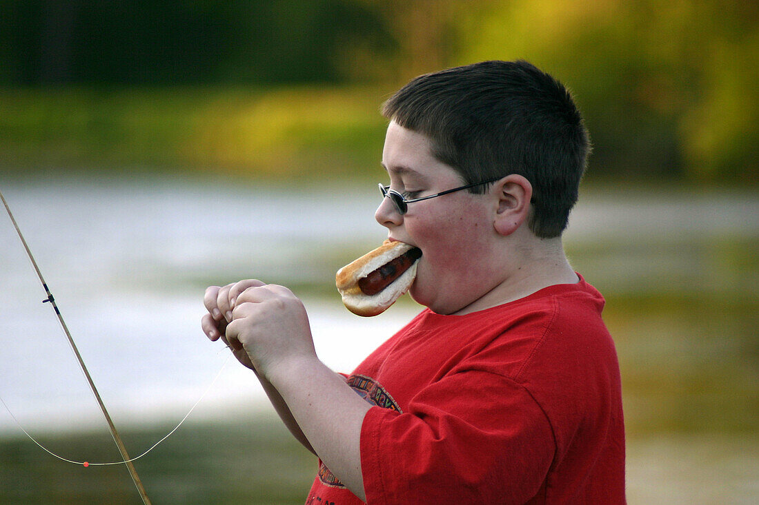 11 year old boy, eating hotdog while baiting hook for fishing