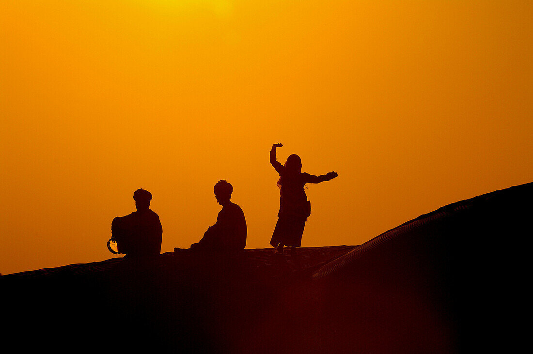 Rajasthani men sit while a woman dances on Kanoi Sand Dunes at sunset, Thar Desert, near Jaisalmer, Rajasthan, India