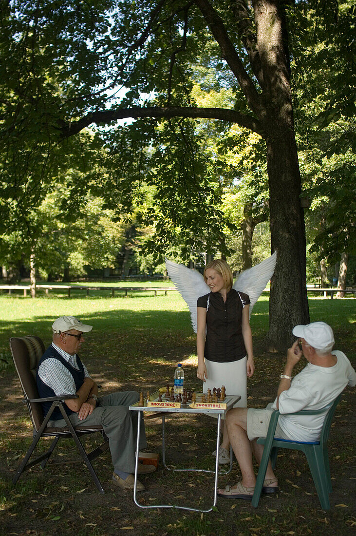 Engel, junge Frau mit Engelsflügeln schaut zu, Zwei ältere Männer spielen Schach im Park, München, Bayern, Deutschland