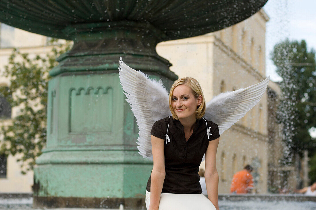 Mid adult woman wearing angel wings sitting near fountain at university, Munich, Bavaria, Germany