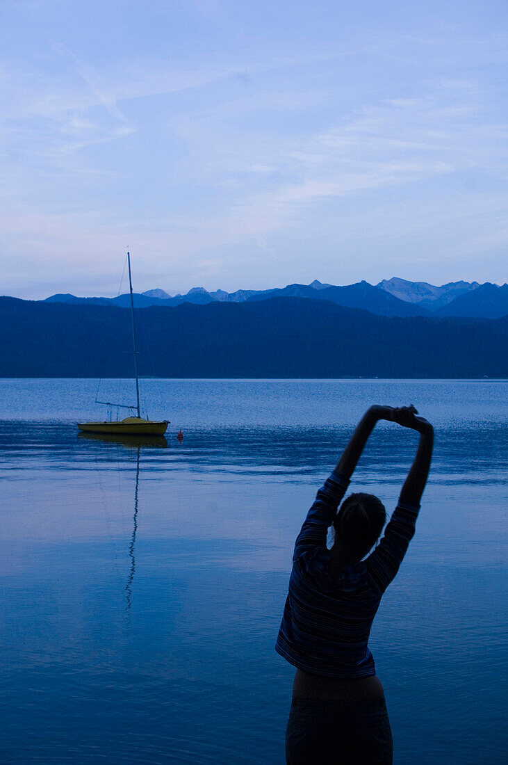 Junge Frau streckt sich am Walchensee in der Abenddämmerung, Bayern, Deutschland