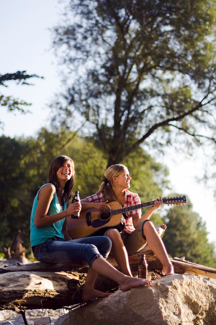 Two young women sitting on the banks of the river Isar in the evening, playing a guitar, Munich, Bavaria, Germany
