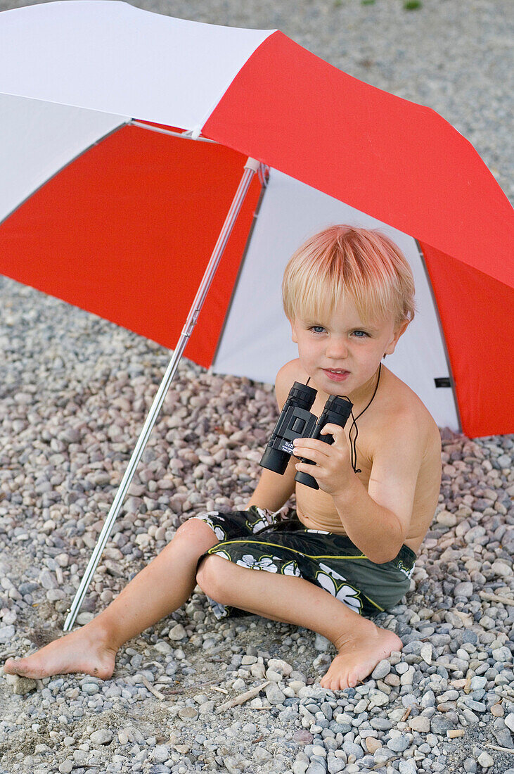 Junge (4-5 Jahre) mit einem Fernglas sitzt unter einem Sonnenschirm