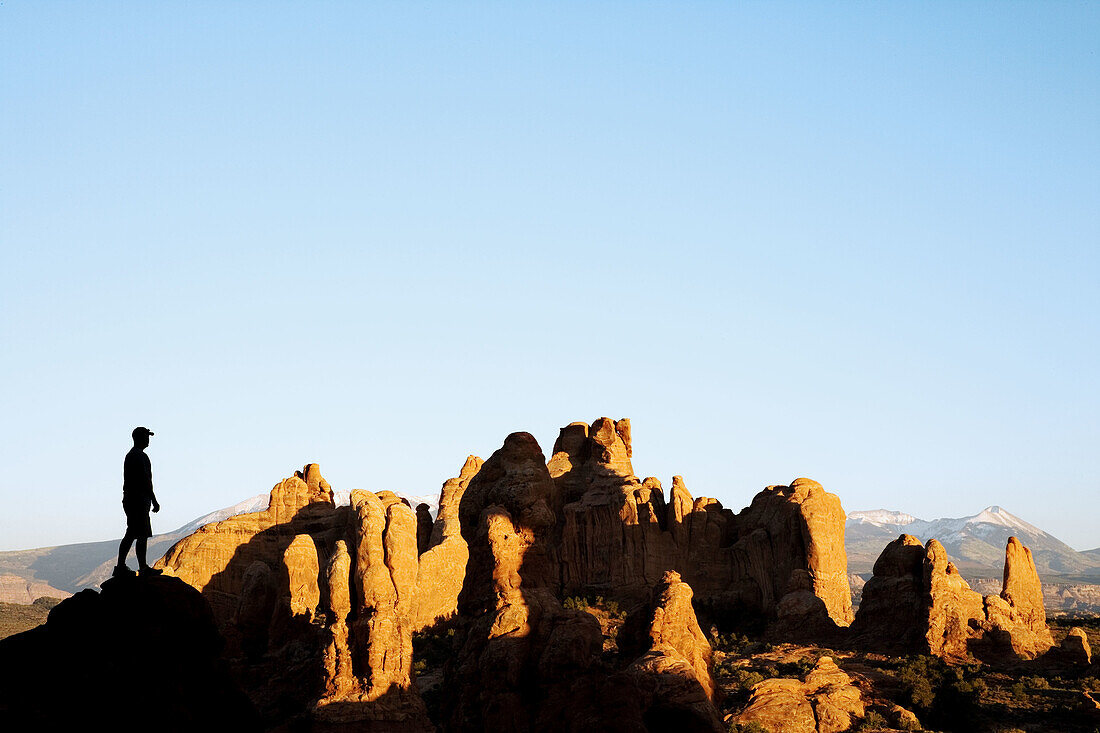 A hiker stops to admire the last rays of light on sandstone fins and snowcovered La Sal mountains, Arches National Park, Utah. USA.