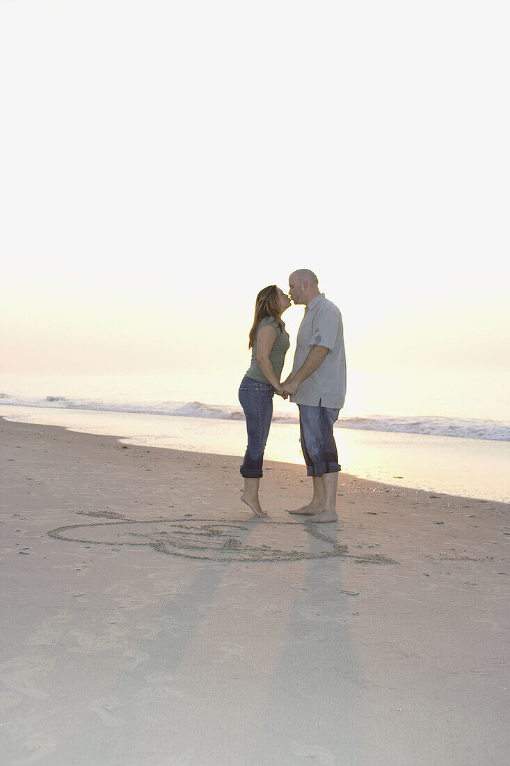 Couple in their 20s kissing on the beach of ocean city by the drawing of heart and arrow and their initials
